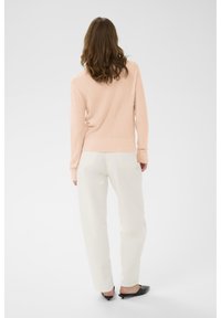 Hellrosa gerippter Pullover mit langen Ärmeln, kombiniert mit weißen Hosen mit geradem Bein und schwarzen Slippern. Einfaches, minimalistisches Design.