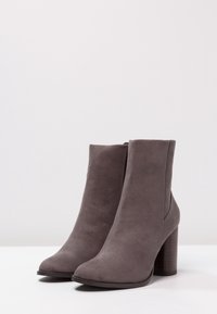 Botas de tobillo de ante gris con punta redondeada y tacón de madera grueso. Textura suave con un diseño elegante y cierre de cremallera en la parte posterior.