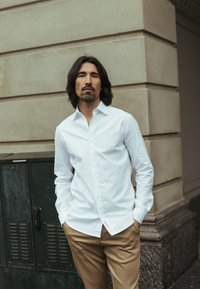 Fehér, hosszú ujjú, gombos ing, világosbarna nadrággal viselve. Sima anyag látható varrási részletekkel, egy texturált fal előtt.