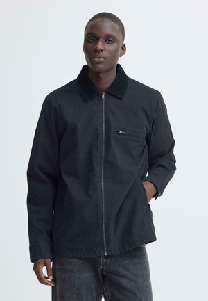 Zwarte man met een donker jack met rits en een ribfluwelen kraag, één hand in de zak, tegen een effen lichtgrijze achtergrond.