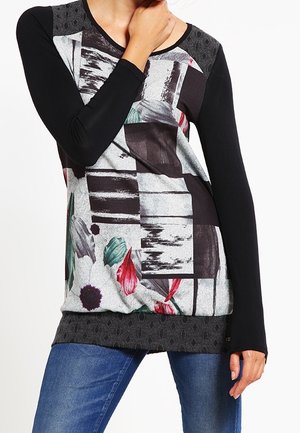 Femme portant un haut noir à manches longues avec des motifs géométriques et floraux abstraits, associée à un jean bleu, main touchant le cou.