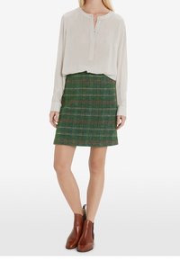 Vaaleanbeige pusero, jossa on pitkät hihat ja henley-kaulus, yhdistetty vihreään ruudulliseen hameeseen sekä ruskeisiin nilkkureihin. Hame ulottuu keskireidestä.