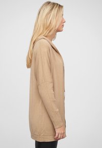 Beige gebreide cardigan met geribde mouwen, open voorkant en een langere achterkant. Zachte textuur en relaxte pasvorm, geschikt voor lagen.
