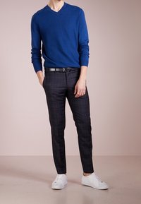 Modrý svetr s výstřihem do V z měkkého materiálu, černé a šedé kostkované kalhoty a bílé tenisky. Minimalistický design bez viditelných vzorů na botách.