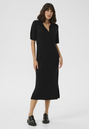 Black polo dress with short sleeves, knee-length cut, side slit detail, soft fabric texture, paired with black loafers.