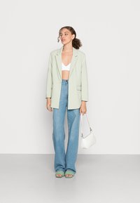 Blazer oversized verde claro, top cropped branco, jeans de cintura alta, mala branca e sandálias verdes de tiras, em modelo contra um fundo neutro.