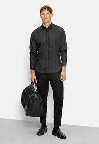 Mann in einem dunkelgrauen Hemd mit Knöpfen, schwarzen Hosen und schwarzen Slippern, der in seiner linken Hand eine große schwarze Lederhandtasche hält.