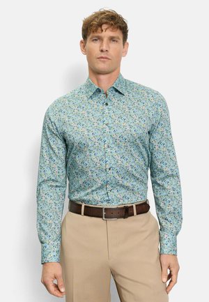 Mann trägt ein eng anliegendes, hellblaues, geblümtes Hemd mit Knöpfen, das in beige Hosen mit einem dunkelbraunen Gürtel gesteckt ist, und steht vor einem schlichten Hintergrund.