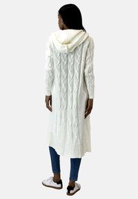 Witte, gebreide lange cardigan met capuchon, voorzien van een kabelbreipatroon en geribde manchetten, gecombineerd met een spijkerbroek en sneakers.