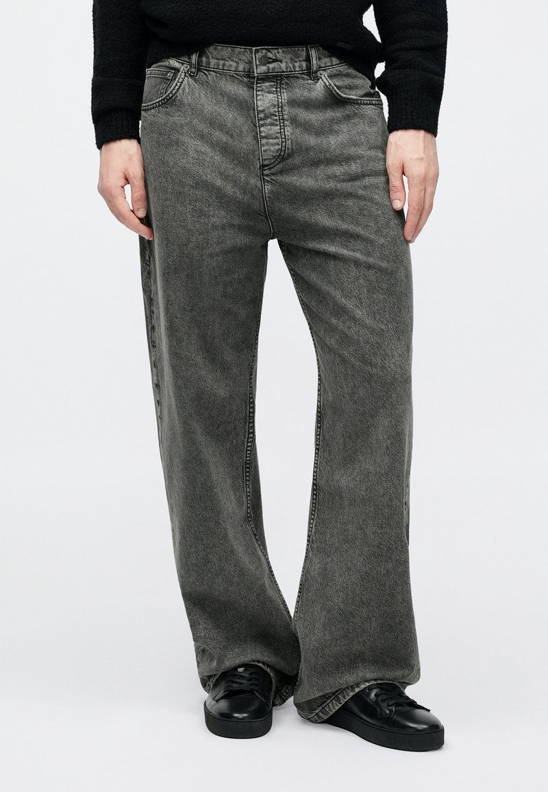 Person wearing loose-fit faded gray denim jeans and black sneakers, standing against a plain white background.