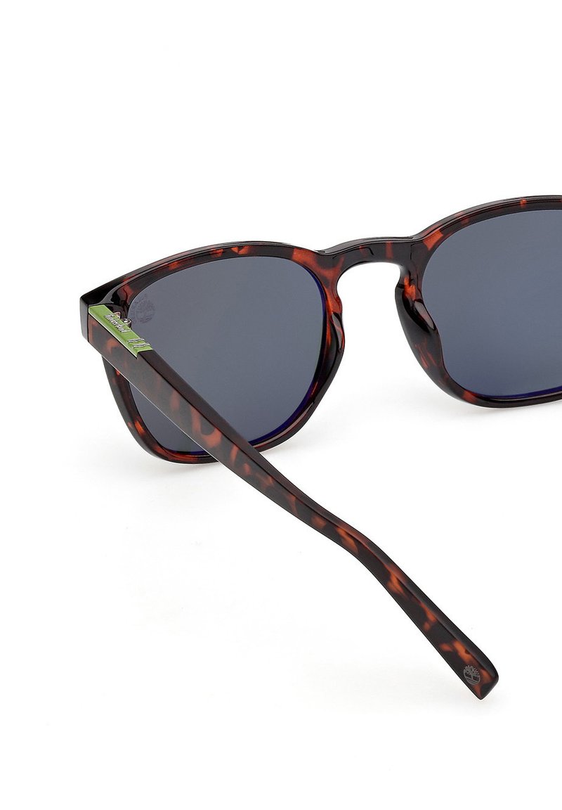 Lunettes de soleil en écaille de tortue avec verres foncés, de forme angulaire et accents verts sur les branches, présentant une monture texturée pour une meilleure prise.