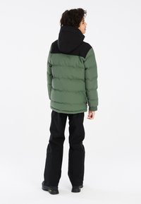 Giacca a vento verde con cappuccio nero, caratterizzata da un design trapuntato, polsini a coste e una vestibilità comoda. Indossata con pantaloni neri e stivali.