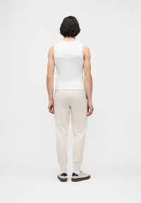 Άτομο με κοντά σκούρα μαλλιά, που φορά λευκό αμάνικο τοπ, ανοιχτό μπεζ παντελόνι jogger και λευκά αθλητικά παπούτσια, στέκεται γυρισμένο με την πλάτη σε απλό φόντο.