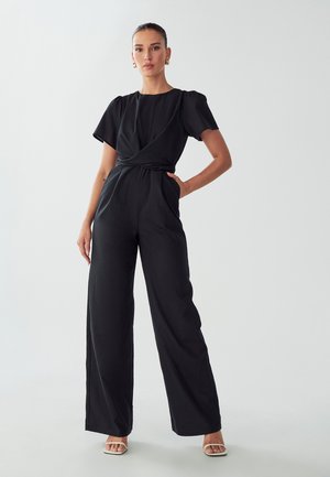 Black wide-leg jumpsuit with short puff sleeves, a wrap design at the waist, and side pockets. Smooth fabric and minimalistic style.