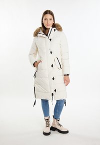 Hvid polstret parka med en faux fur hætte, sorte accenter, sidelommer og justerbare snore, parret med lyseblå jeans og beige støvler.