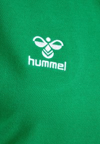 Hvidt broderet bi-logo og "hummel" tekst på grøn tekstureret stof, sandsynligvis sportstøj eller atletisk beklædning.