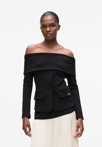 Black off-the-shoulder jacket with long sleeves, single-button closure, and two front pockets, paired with a pleated white skirt.