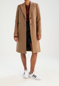 Cappotto di lana beige dal taglio classico, con rever a punta e due bottoni, abbinato a una gonna di pelle nera e sneakers bianche.