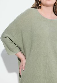 Maglione verde oversize lavorato a maglia con un motivo testurizzato, ampia scollatura a barca e maniche morbide. Mano parzialmente visibile nella tasca.