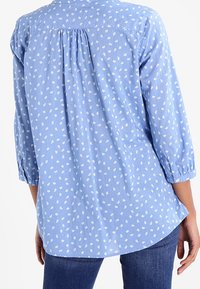 Chemise bleu clair à manches longues avec imprimé de feuilles blanches, détail de pli au dos et ourlet incurvé. Fabriquée en tissu léger.