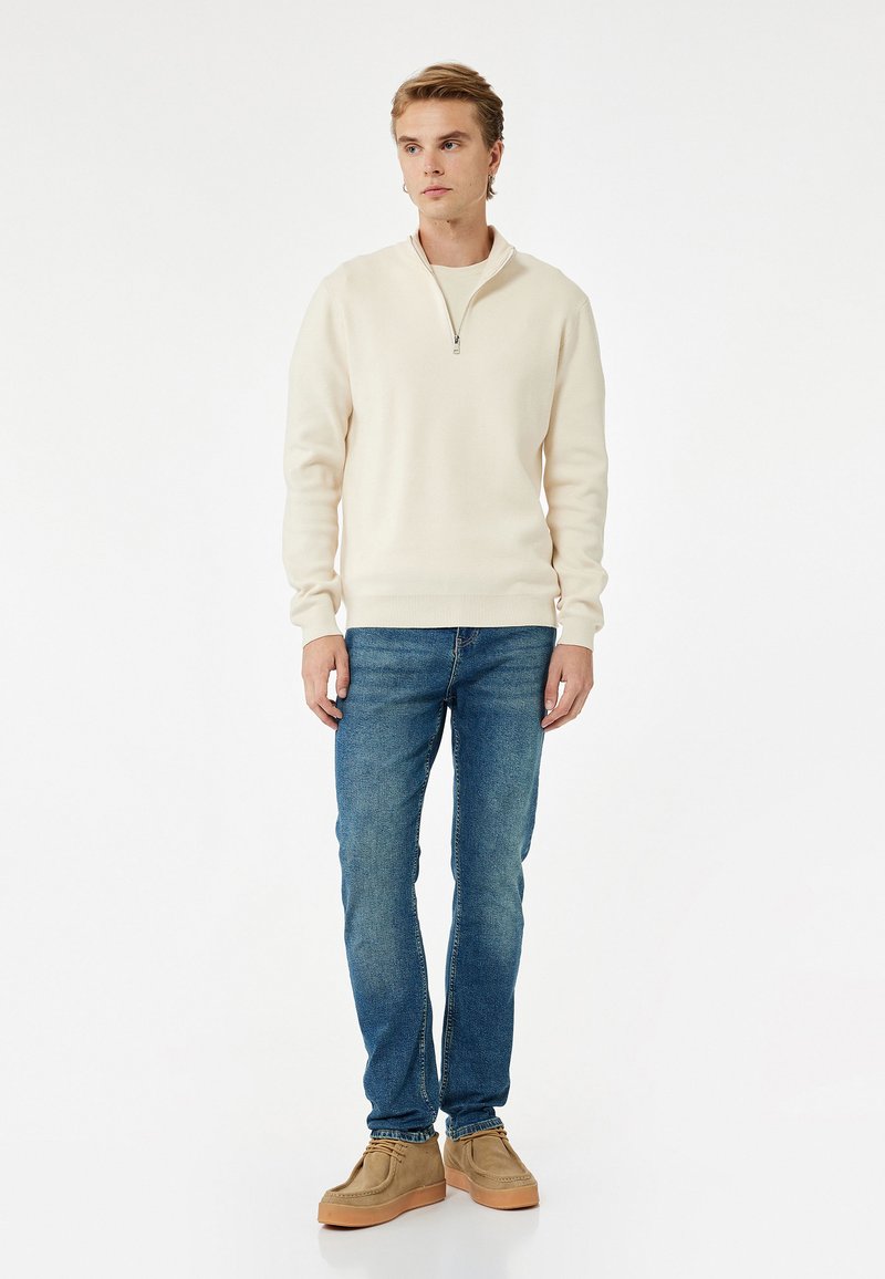 Suéter de color crema claro con cuello con cremallera, combinado con jeans azules y zapatos beige. Textura suave, diseño liso y ajuste relajado.