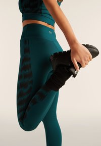 Leggings deportivos en color verde azulado con un patrón de zigzag negro, combinados con unas zapatillas negras que tienen una superficie texturizada y detalle acanalado en el tobillo.