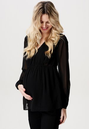 Black maternity blouse with a V-neck, long sheer sleeves, and a gathered waist, styled over black pants, worn comfortably over a rounded belly.