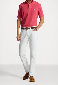 Röd polo skjorta, vita jeans, brunt läderbälte och bruna mockaskor. Anteckningsvärda detaljer inkluderar en liten logotyp på skjortan och ett armbandsur.