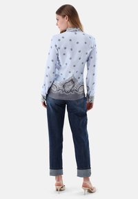 Blusa celeste con motivo floreale nero, maniche lunghe e orlo decorativo a paisley, abbinata a jeans scuri arrotolati e tacchi beige.