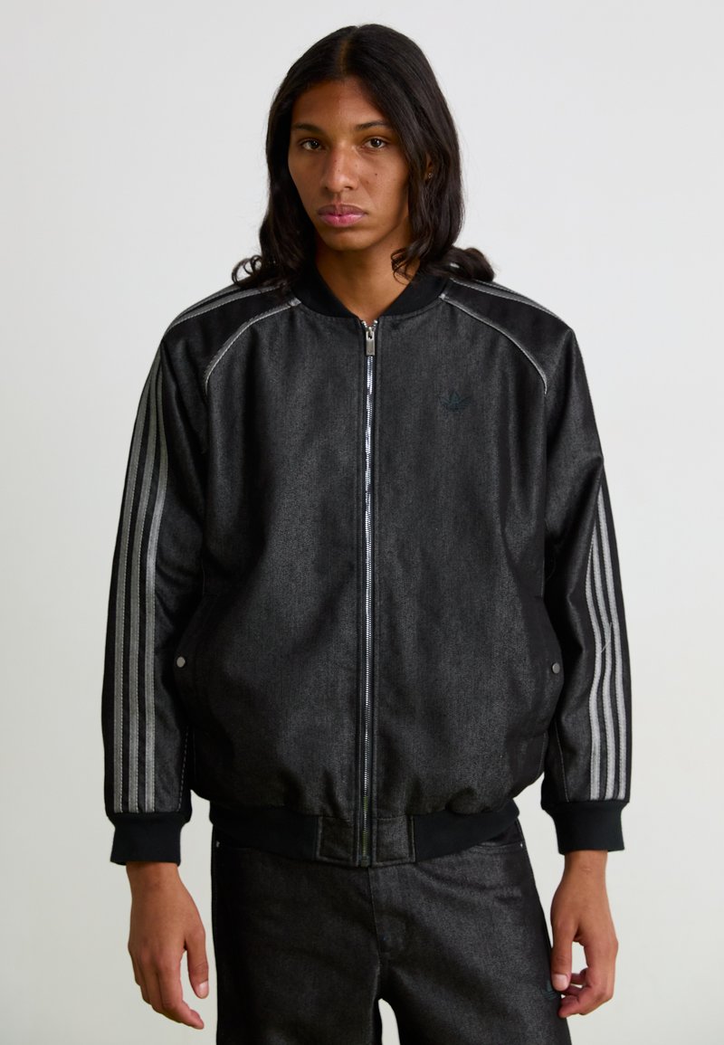 Chaqueta bomber negra con un acabado texturizado, rayas plateadas en las mangas, cierre frontal con cremallera y puños de canalé. Pantalones a juego mostrados.