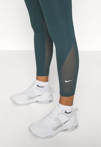 Zielone legginsy z siateczkowymi panelami w połączeniu z białymi trampkami Nike z srebrnymi akcentami i czarnym logo. Tekstura jest gładka i dopasowana.