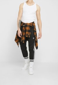 Svarta träningsbyxor med vita ränder, en orange och svart rutiga skjorta knuten i midjan, och en vit linne. Vita sneakers.