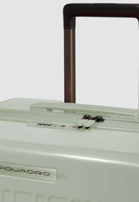 Valise rigide vert clair avec une finition lisse, fermeture éclair, doubles curseurs noirs et poignée rétractable marron foncé. Logo Piquadro affiché.