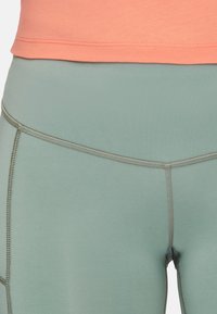 Leggings leggeri di colore verde menta con vita alta, caratterizzati da cuciture piatte e una texture liscia, abbinati a un top corallo appena sopra la vita.