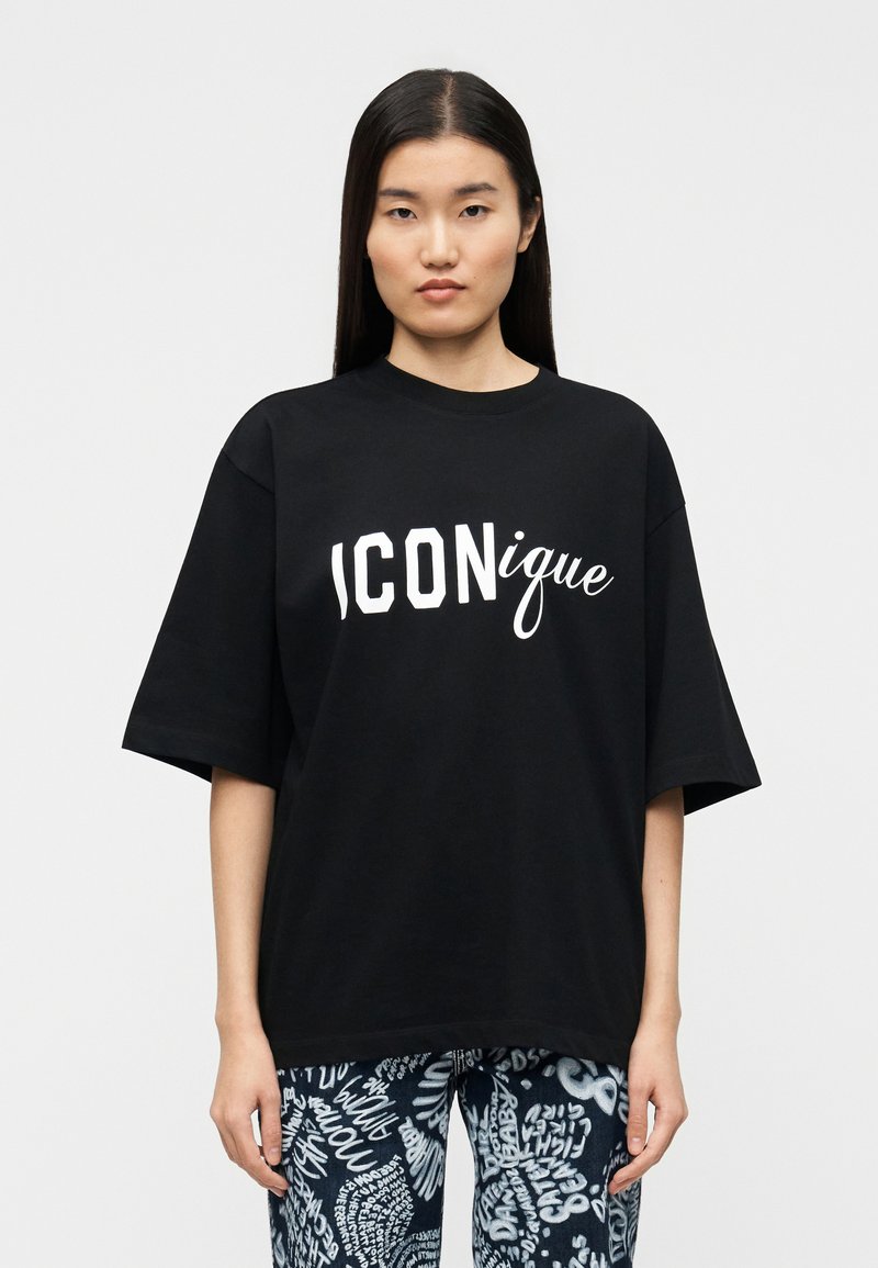 Μαύρο βαμβακερό T-shirt σε υπερμεγέθη γραμμή με λευκό κείμενο "ICONique", κοντά μανίκια, στρογγυλή λαιμόκοψη και άνετη εφαρμογή, συνδυασμένο με μπλε τζιν με σχέδια.