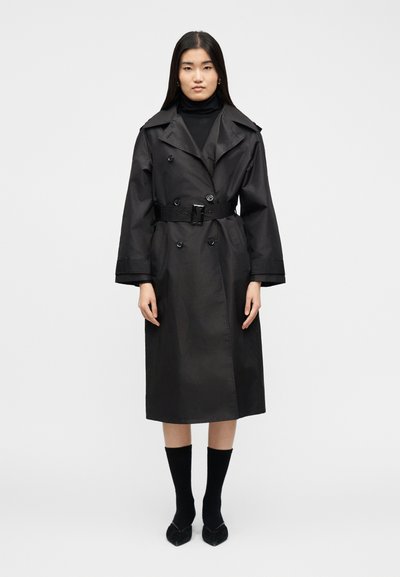 Une femme debout face à l'avant portant un trench-coat noir ceinturé sur un col roulé noir, avec des chaussettes noires et des chaussures pointues sur un fond blanc.