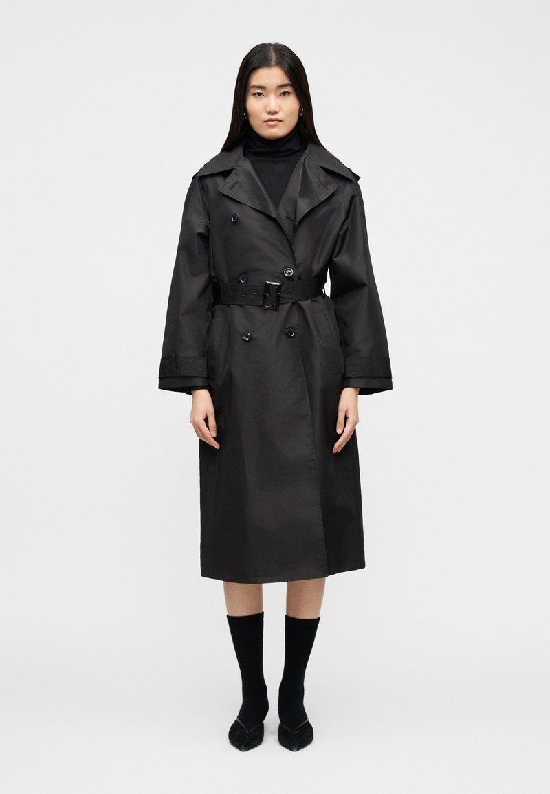 Γυναίκα στέκεται με το σώμα στραμμένο προς τα εμπρός, φορώντας ένα μαύρο παλτό τύπου trench με ζώνη πάνω από μαύρο ζιβάγκο, μαύρες κάλτσες και μυτερές παπούτσια, σε λευκό φόντο.