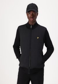 Black vest with quilted front and ribbed knit sleeves. Features a high collar and zip closure. A small yellow logo is displayed on the chest.