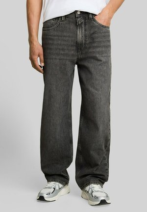 Mann trägt lockere dunkelgraue Jeans und weiße Sneakers mit schwarzen und grauen Details, steht mit einer Hand in der Tasche.