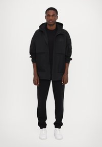 Svart huva jacka med mesh-fickor, kombinerad med svarta byxor och vita sneakers. Tyget verkar lättviktigt och texturerat med en avslappnad passform.