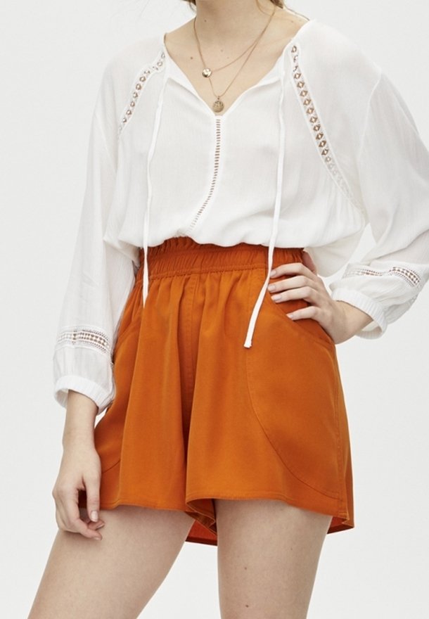 Femme portant un chemisier blanc à manches longues avec des détails en dentelle et un short taille haute orange brûlé avec des poches, main sur la hanche.