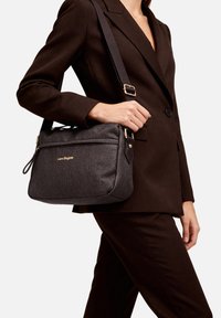 Persona con indosso un completo marrone scuro che tiene una borsa a tracolla grigio scuro con trama, logo dorato e tracolla regolabile sulla spalla.