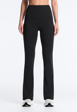 Persoană purtând colanți negri evazați, mulați, cu talie înaltă și pantofi sport albi, stând pe un fundal alb simplu.