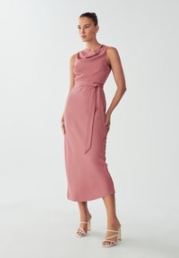 Sleeveless, knee-length dress in soft dusty rose fabric, featuring a draped neckline and a tied waist sash; paired with white strappy heels.