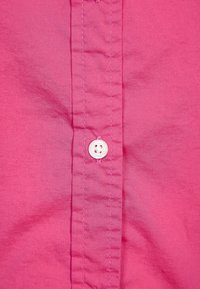 Rosa Hemd mit Knopf, das einen weißen Knopf, eine ordentlich vernähte Knopfleiste und eine glatte Textur aufweist. Der Stoff hat einen leichten Glanz.