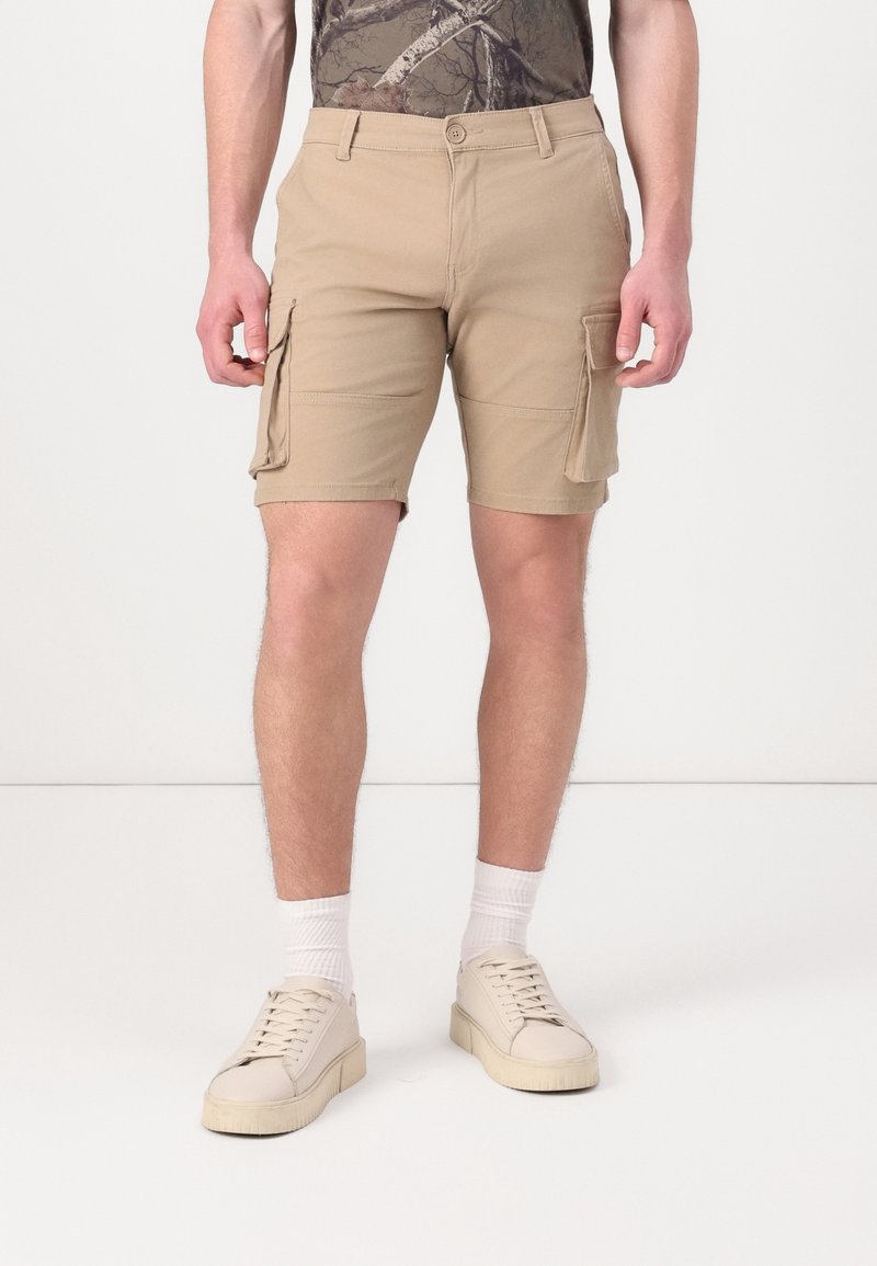 Shorts cargo en kaki en mélange de coton, dotés de poches latérales, d'une coupe droite et d'une fermeture à bouton, associés à des baskets blanches et des chaussettes.