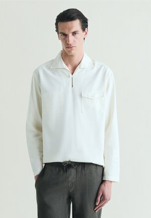 Hombre con el pelo peinado hacia atrás lleva una camisa blanca de manga larga con un bolsillo delantero y pantalones oscuros con cordón, de pie contra un fondo liso.