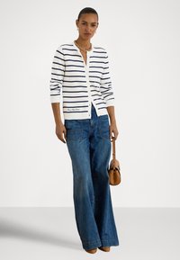 Λευκή ζακέτα με ναυτικό μπλε ρίγες, κλείσιμο με κουμπιά, ριμπ μανσέτες, συνδυασμένη με παντελόνι τζιν με ευρύ πόδι σε μπλε απόχρωση. Μικρή καφέ τσάντα χιαστί ως αξεσουάρ.