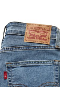 Jeans de denim azul con un parche de cuero marrón que lleva un logo y gráficos en relieve en la cintura. Etiqueta roja visible en la costura lateral.