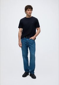 Jeune homme debout portant un t-shirt noir, un jean bleu, une ceinture noire et des chaussures noires sur un fond clair uni.